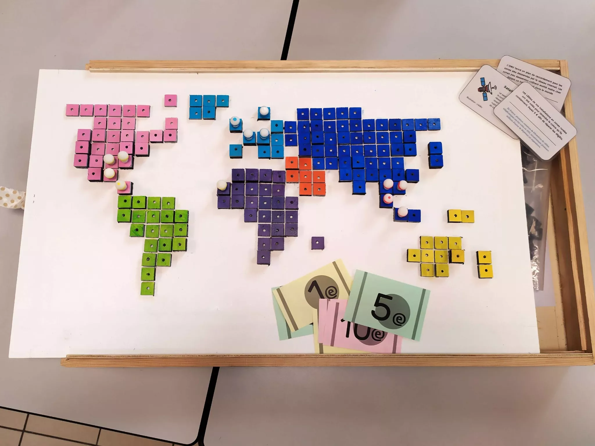 Une photo du jeu fini. On y voit le plateau de jeu avec des continents faits de tuiles carrées et peintes de différentes couleurs. Des pions sont placés sur certaines tuiles et des cartes et faux billets sont sortis de la boîte entrouverte.